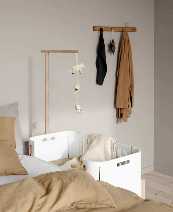 Oliver Furniture witte en eikenhouten co-sleeper naast een volwassen bed, omgezet naar een kinderbankje in een huiselijke setting.