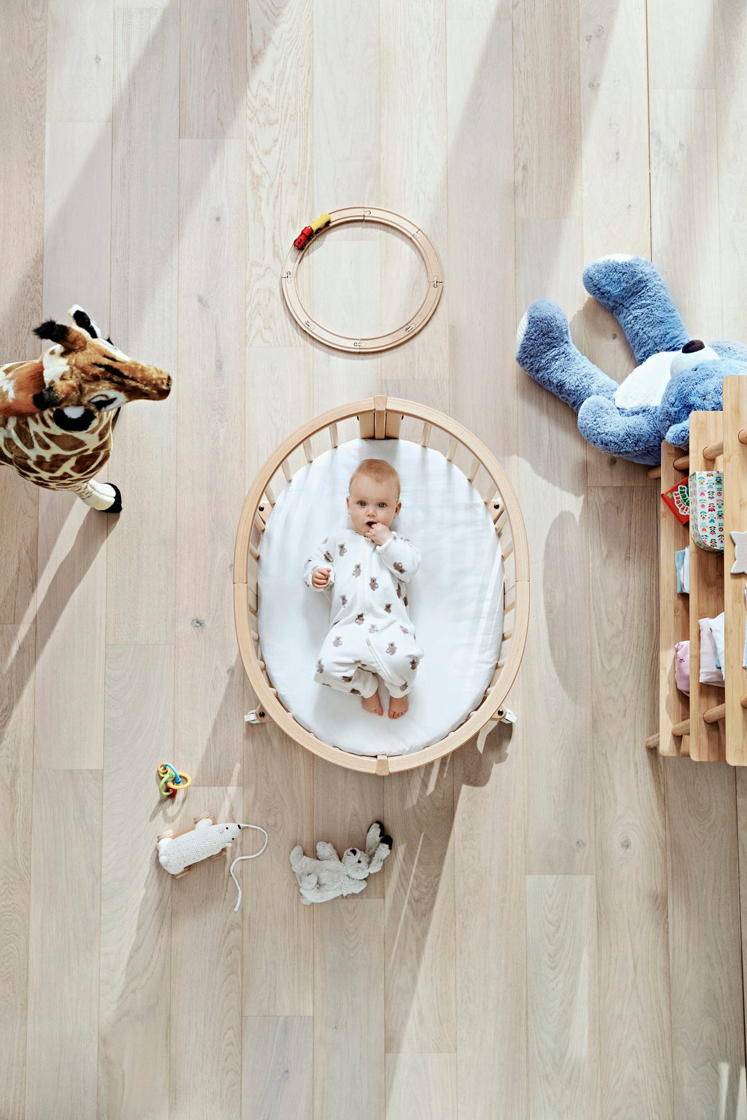 Stokke Sleepi™ Mini V3 wieg in natuurlijke houtafwerking, met zachte ovale vorm en vergrendelbare zwenkwielen.